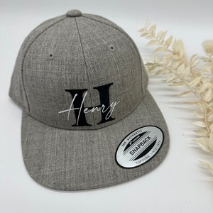 Könnte beinhalten: Eine graue Baseballkappe mit einem schwarzen und weißen gestickten Buchstaben "H" und dem Namen "Henry" in Kursivschrift. Die Kappe hat einen schwarzen verstellbaren Druckknopfverschluss mit einem silbernen Etikett, auf dem "The Authentic Yupoong Snapback Since 1974" steht.