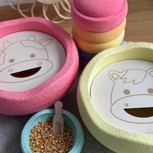 Könnte beinhalten: Ein Set aus bunten Stapelbechern mit einem Kuh-Design. Die Becher sind aus Schaumstoff und haben eine strukturierte Oberfläche. Die Becher sind in Regenbogenfarben gestapelt. Im Vordergrund befindet sich eine kleine Schüssel mit Maiskörnern und ein Holzlöffel.