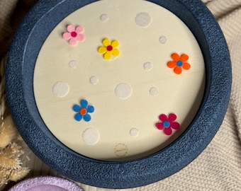 May include: A navy blue tray with a light wood interior decorated with colorful felt flowers and white polka dots. A smaller purple tray with felt flowers is in the foreground. The background includes a woven circular object and a beige fabric.