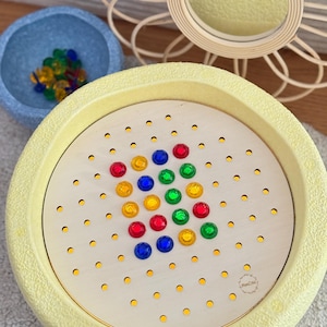 Könnte beinhalten: Eine gelbe Plastikschale mit einem Holzkreis im Inneren. Der Kreis hat ein Gitter aus Löchern, in die farbige Plastikpfähle in einem Muster gesteckt werden. Die Pfähle sind rot, blau, grün und gelb. Die Schale hat ein weißes Etikett mit dem Text "Wood City" darauf.