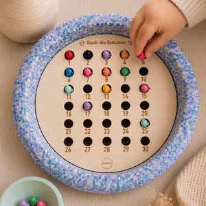 Könnte beinhalten: Ein kreisförmiges Spielbrett mit einem blau-violett gesprenkelten Rand und einer hellen Holzmitte mit dreißig nummerierten Löchern. Kleine bunte Entenfiguren stecken in einigen Löchern, mit dem aufgedruckten Text Such die Entchen oben.
