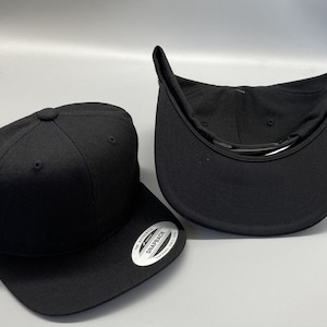 May include: Two black baseball caps, one with the inside facing up and the other with the outside facing up. The cap with the inside facing up has a black strap and the text "The Authentic Snapback" printed on the inside of the cap.