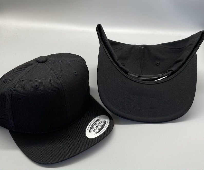 May include: Two black baseball caps, one with the inside facing up and the other with the outside facing up. The cap with the inside facing up has a black strap and the text "The Authentic Snapback" printed on the inside of the cap.