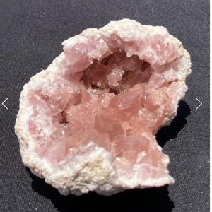 Puede incluir: Geoda de cristal rosa con un exterior rugoso y blanco. El interior revela un grupo de cristales rosas, creando una estructura similar a una cueva. La geoda se encuentra sobre un fondo oscuro, resaltando su belleza natural.