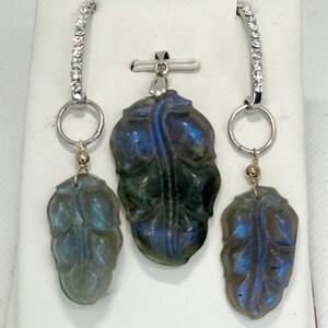 Puede incluir: Un conjunto de joyas con un colgante y pendientes. El colgante y los pendientes tienen un diseño de hoja tallada en una piedra azul-verde. Los pendientes tienen un aro plateado con piedras transparentes.