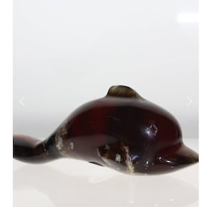 Puede incluir: Figurita de delfín marrón oscuro brillante, con un acabado liso y pulido. El delfín tiene un cuerpo curvado y una pequeña aleta dorsal. La figura tiene marcas blancas y marrón claro en el costado. El delfín está sobre una superficie reflectante.