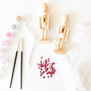 Puede incluir: Dos figuras de cascanueces de madera, botes de pintura, pinceles y lentejuelas rojas y plateadas sobre una superficie blanca.