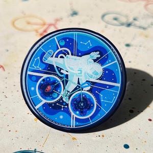 Op de afbeelding: Een blauwe en witte emaille pin met een astronaut die op een fiets door de ruimte fietst. De astronaut draagt een wit ruimtepak en een helm. De fiets heeft een wit frame en zwarte banden. De achtergrond is donkerblauw met witte sterren en planeten.