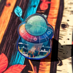 May include: A colorful enamel pin featuring a blue and green spaceship with a forest scene inside. The pin is set against a woodgrain background.
