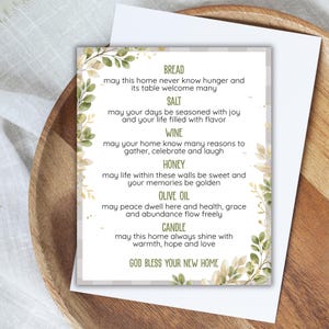 May include: A rectangular housewarming card featuring a white background with a light gray plaid border and green leaf illustrations. The text lists blessings for bread, salt, wine, honey, olive oil, and a candle, ending with God bless your new home.