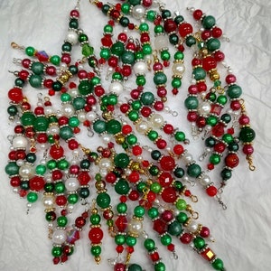 May include: A collection of Christmas ornaments with red, green, white, and gold beads. The ornaments are strung together with wire and feature various shapes and sizes of beads, including pearls and faceted crystals. The ornaments are arranged on a white surface.