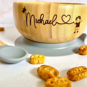 bamboo bowl with a personalised baby name, the robust silicone suction base helps the bowl stick on the flat surface. Next to it is a bamboo spoon with grey food-grade non-toxin and BPA silicone