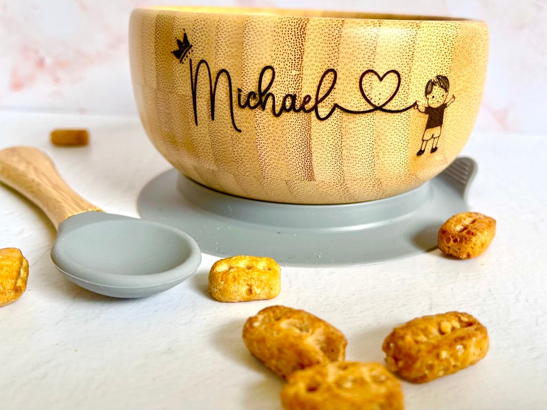 bamboo bowl with a personalised baby name, the robust silicone suction base helps the bowl stick on the flat surface. Next to it is a bamboo spoon with grey food-grade non-toxin and BPA silicone