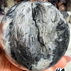 May include: A spherical black and white mineral specimen featuring a prominent vertical fissure and fibrous, needle-like crystalline textures. The surface displays a mix of opaque black sections and translucent white areas with a polished finish.