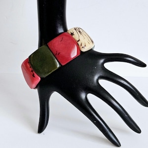 May include: A colourful square-shaped bracelet made of red, green and cream coloured plastic. The bracelet is on a black hand model.