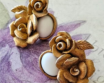 May include: A pair of stud earrings featuring a metallic gold-tone finish with sculpted rose motifs and a smooth, round white cabochon center. The floral design includes detailed petals and leaves, creating a dimensional appearance for each earring.