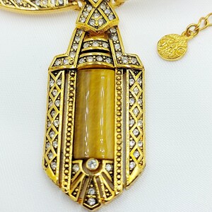May include: Gold-toned necklace with a pendant featuring a rectangular, tiger's eye-colored stone. The pendant is framed with clear crystals and ornate gold detailing. The necklace has a matching chain and clasp.