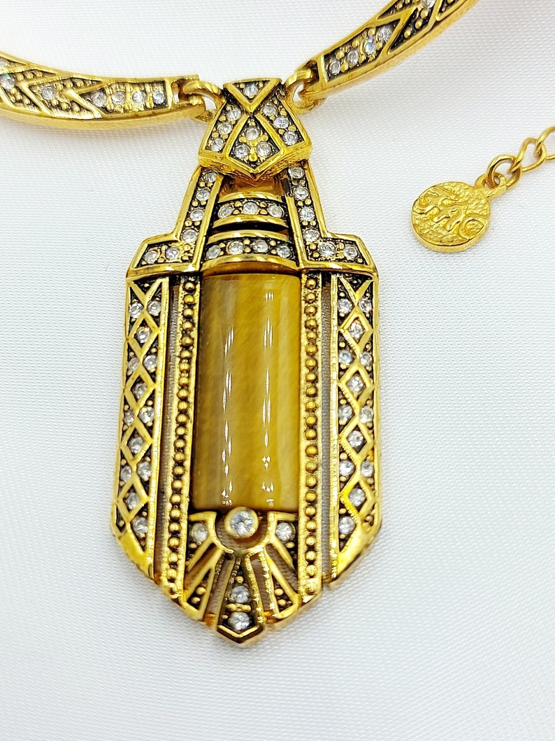 May include: Gold-toned necklace with a pendant featuring a rectangular, tiger's eye-colored stone. The pendant is framed with clear crystals and ornate gold detailing. The necklace has a matching chain and clasp.