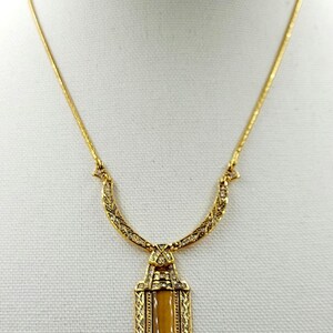 May include: Gold-toned necklace with a rectangular pendant featuring a light brown center stone. The pendant and chain are adorned with intricate detailing and small, clear embellishments. The necklace is displayed on a white surface.