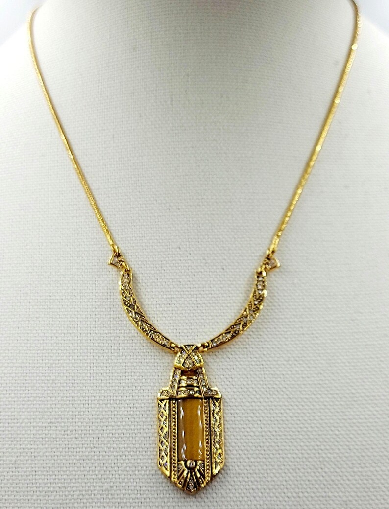 May include: Gold-toned necklace with a rectangular pendant featuring a light brown center stone. The pendant and chain are adorned with intricate detailing and small, clear embellishments. The necklace is displayed on a white surface.