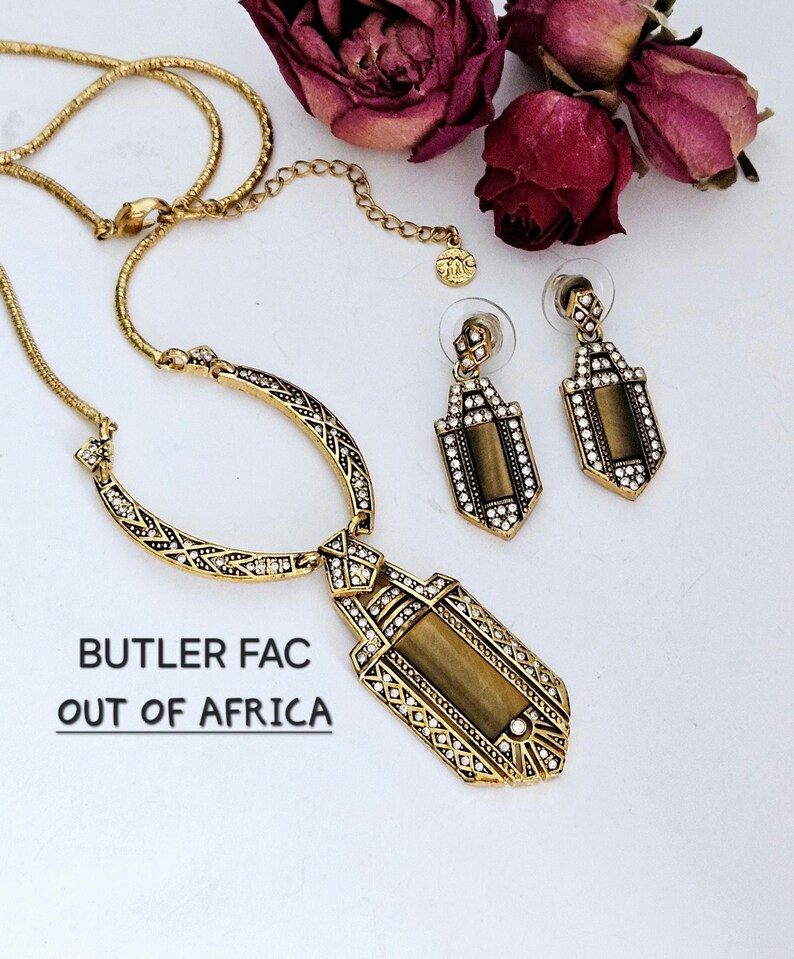 May include: A gold-toned necklace and matching earrings set with a geometric design. The necklace features a pendant with a rectangular center and is embellished with clear crystals. The earrings have a similar design. The text "BUTLER FAC OUT OF AFRICA" is visible.
