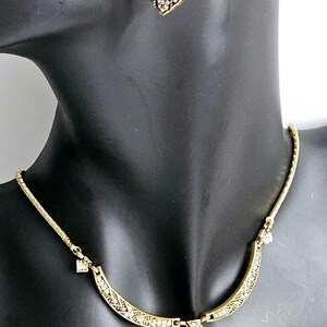 May include: Gold-toned necklace and earring set. The necklace features a pendant with a rectangular amber-colored stone, surrounded by clear crystals. The matching earrings have a similar design. The jewelry is displayed on a black mannequin.