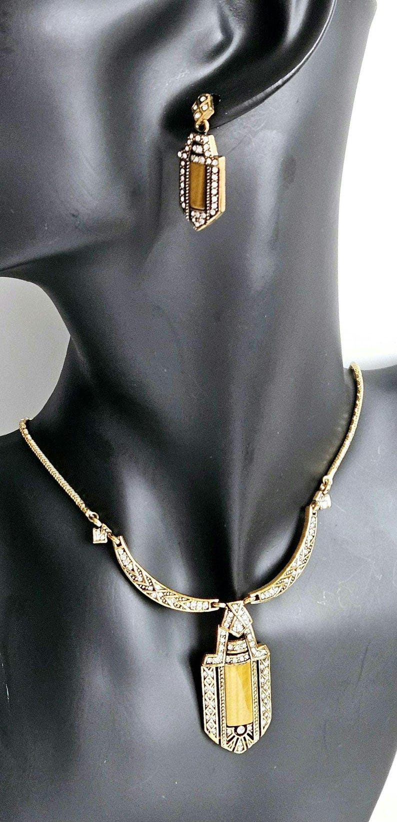 May include: Gold-toned necklace and earring set. The necklace features a pendant with a rectangular amber-colored stone, surrounded by clear crystals. The matching earrings have a similar design. The jewelry is displayed on a black mannequin.