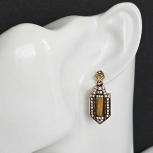 May include: A gold-toned earring with a rectangular pendant. The pendant is outlined with small, clear crystals and features a central, rectangular, amber-colored stone. The earring is displayed on a white surface.