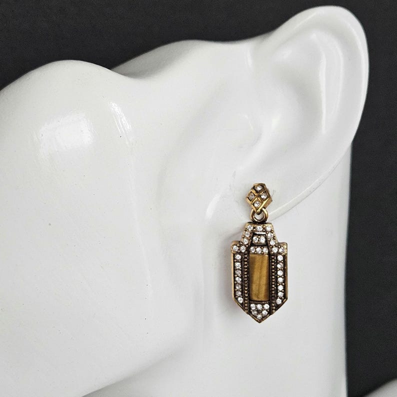 May include: A gold-toned earring with a rectangular pendant. The pendant is outlined with small, clear crystals and features a central, rectangular, amber-colored stone. The earring is displayed on a white surface.