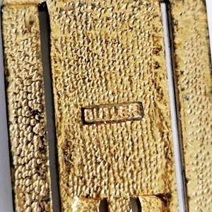 May include: Close-up of a gold-toned metal object with a textured surface. The word "BUTLER" is stamped on a small rectangular plate. The object has a rectangular shape with a vertical design.