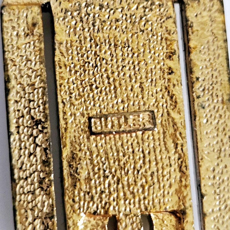 May include: Close-up of a gold-toned metal object with a textured surface. The word "BUTLER" is stamped on a small rectangular plate. The object has a rectangular shape with a vertical design.