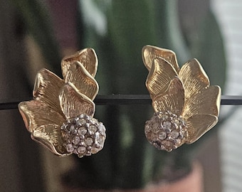 May include: A pair of gold-toned earrings featuring a textured, layered petal design that fans out from a central, spherical cluster of small, clear, sparkling crystals. The metal surface has a brushed finish with visible fine lines.