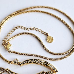 May include: A gold-toned necklace with a delicate chain and an ornate pendant. The pendant features a geometric design with embedded clear crystals. The necklace is displayed on a white surface, highlighting its intricate details and elegant style.