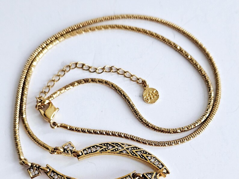 May include: A gold-toned necklace with a delicate chain and an ornate pendant. The pendant features a geometric design with embedded clear crystals. The necklace is displayed on a white surface, highlighting its intricate details and elegant style.