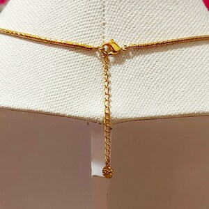 May include: Gold-colored necklace with a delicate chain and a lobster clasp. The necklace is displayed on a white surface, showcasing the intricate details of the jewelry. The extender chain adds adjustability.