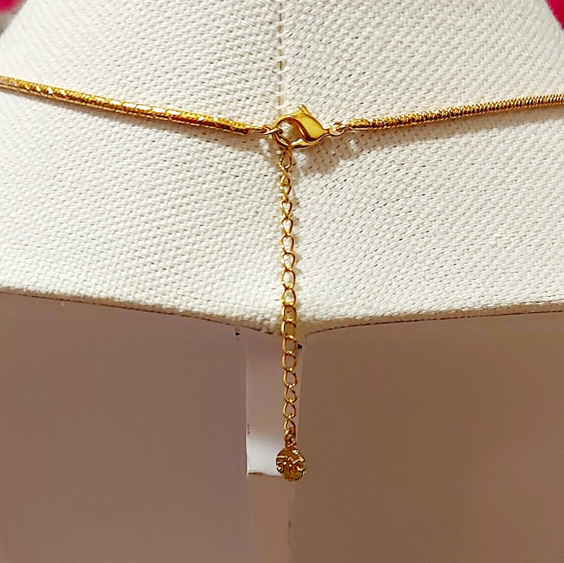 May include: Gold-colored necklace with a delicate chain and a lobster clasp. The necklace is displayed on a white surface, showcasing the intricate details of the jewelry. The extender chain adds adjustability.