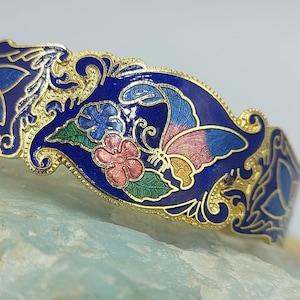 May include: A gold-toned bracelet with blue enamel and a butterfly design. The butterfly has pink, blue, and yellow wings. The bracelet is decorated with floral motifs.