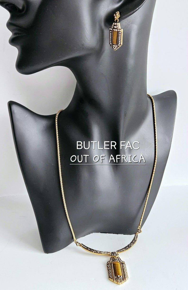 May include: Gold-toned necklace and matching earrings set displayed on a black mannequin. The jewelry features rectangular pendants with a brown center stone and ornate detailing. The text "BUTLER FAC OUT OF AFRICA" is visible.