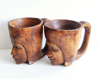 May include: Two wooden mugs carved in the shape of human faces. The mugs are brown and have a rustic appearance.