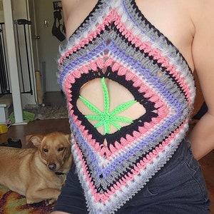 Puede incluir: Un top halter tejido a crochet con un patrón de rayas de colores y un recorte de hoja de marihuana verde en el centro. El top se lleva sobre unos pantalones cortos negros.