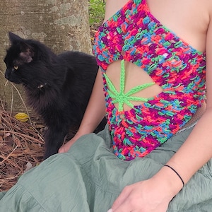 Puede incluir: Una mujer lleva un top de crochet colorido con un diseño de hoja de marihuana verde. Está sentada en el suelo con un gato negro a su lado.