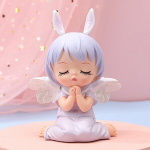 Peut inclure: Une petite figurine d'ange en couleur violette avec des ailes blanches et des oreilles de lapin. L'ange est assis les mains jointes en prière et les yeux fermés.