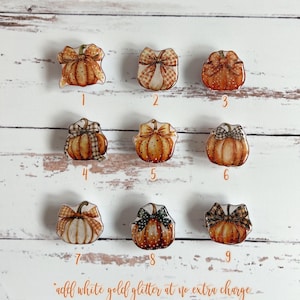 Könnte beinhalten: Neun verschiedene Kürbis-X-Ray-Markierer-Knöpfe. Jeder Knopf zeigt ein anderes Design mit einem Kürbis, einer Schleife und verschiedenen Mustern. Die Knöpfe sind von 1 bis 9 nummeriert. Der Text "add white gold glitter at no extra charge" befindet sich am unteren Rand des Bildes.
