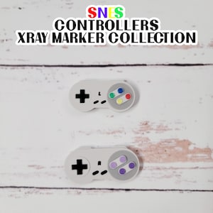 May include: Two white SNES controllers with different colored buttons. The top controller has yellow, green, red, and blue buttons. The bottom controller has purple buttons. The controllers are on a white wooden background. SNES CONTROLLERS XRAY MARKER COLLECTION