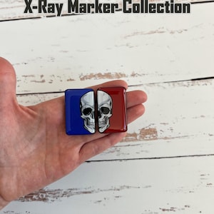 May include: A pair of square markers, one blue and one red, each featuring half of a skull graphic. The markers are held in a hand against a white wood background. The text "Split Skull X-Ray Marker-Collection" is at the top.