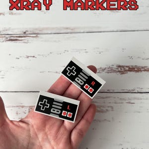 Może przedstawiać: Dwa białe i czarne naklejki kontrolera Nintendo Entertainment System (NES) z czerwonymi przyciskami. Naklejki mają nadrukowane słowa "L" i "R". Naklejki są trzymane w dłoni na tle białego drewna.