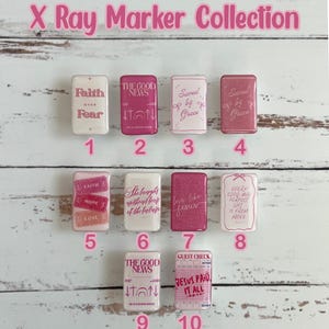 May include: A collection of ten X-ray markers with inspirational messages in pink and white. The markers feature phrases like "Faith over Fear," "The Good News," and "Saved by Grace." The markers are arranged on a light wood-grain surface.