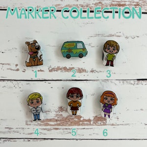 May include: A set of six Scooby Doo themed character markers. The markers are round and feature different characters from the show, including Scooby Doo, Shaggy, Velma, Daphne, Fred, and the Mystery Machine. The markers are numbered 1 through 6.