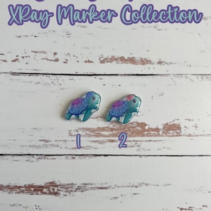 May include: Two sea turtle shaped X-ray markers with purple flowers on their heads. The markers are on a white wooden background. The text "Cutest Sea Turtles X-ray Marker Collection" is at the top of the image. The text "add pixie glitter, no extra charge" is at the bottom of the image.