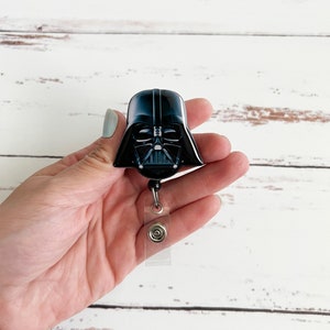 May include: A black and glossy Darth Vader helmet badge reel featuring the iconic character's mask with silver accents. The badge reel includes a retractable cord and a clear plastic strap with a metal snap button for attaching identification cards.